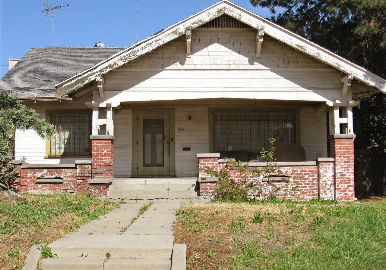 An older Craftsman bungalow in Los Angeles with peeling paint, worn trim, barred windows and a patchy dry front lawn